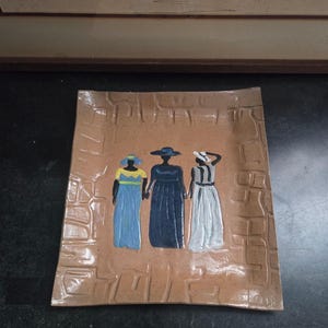May include: A rectangular ceramic plate with a textured brown surface. The plate features a painted image of three figures in long dresses and hats, holding hands. The figures are in blue, black, and white. The plate has a slightly curved shape.
