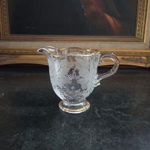 May include: Clear glass creamer with etched floral design and gold trim. The creamer has a handle and a spout. The base is round and also has gold trim. The creamer is likely vintage and is a decorative item.
