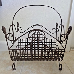 Pode incluir: Um porta-revistas decorativo em ferro forjado com acabamento castanho escuro. O porta-revistas apresenta uma pega curva, detalhes enrolados e uma frente em estilo treliça. O porta-revistas tem um design semelhante a uma cesta com quatro pés curvos.