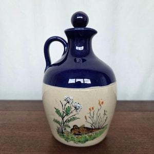 Puede incluir: Jarra de cerámica con tapa azul oscuro y base beige. La base presenta una escena pintada a mano de un faisán en una zona herbosa con flores silvestres y bayas naranjas. La jarra tiene un asa y un tapón redondo.