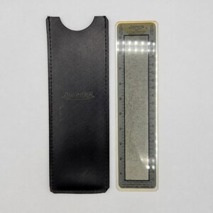 Puede incluir: Una funda protectora negra similar al cuero con la palabra Linometer grabada en el frente, colocada junto a un medidor Linometer rectangular. El medidor presenta un centro metálico con escalas de medición impresas que van de 35 a 80.