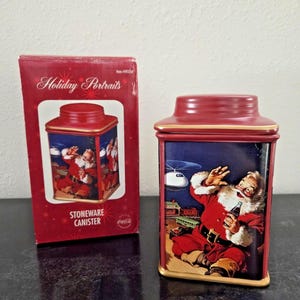 Puede incluir: Un recipiente de gres rojo y su caja a juego con una ilustración de Papá Noel. El recipiente tiene forma rectangular con tapa, y la caja dice "Holiday Portraits" y "Stoneware Canister".