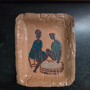 Op de afbeelding: Een rechthoekige, bruinbeige keramische schaal met een opstaande rand en een reliëf ontwerp. De schaal toont een afbeelding van twee figuren in teal en zwarte kleding, zittend bij een mand. De figuren lijken te werken met de inhoud van de mand.