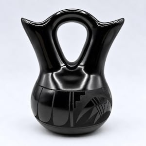 May include: A polished black ceramic vessel featuring a double-spout design connected by a central arched handle. The rounded base is decorated with matte black geometric patterns, including vertical lines and stepped motifs.