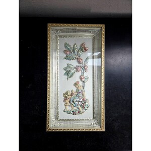 May include: Framed relief art featuring a tree with red berries and figures. The artwork is set in a rectangular frame with a silver and gold border. The figures are dressed in colorful clothing.
