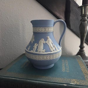 May include: A light blue ceramic pitcher with white relief figures and decorative bands. The pitcher has a handle and is sitting on a stack of antique books with gold lettering.