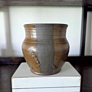 May include: A handcrafted ceramic pot with a brown and gray color scheme. The pot has a textured surface with a vertical gray stripe and embossed circular designs. It sits on a white rectangular base, showcasing its rustic charm.