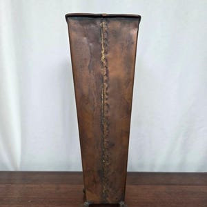 May include: A tall, rectangular copper umbrella stand with a slightly tapered design. The stand has a riveted seam down the center and rests on three small, ball-shaped feet. The copper has a rich, aged patina.