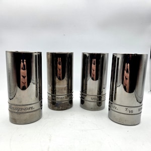 May include: Four cylindrical chrome-finished metal sockets arranged in a row. Each socket features the engraved text Snap-on, SFS141, USA, and 7/16. The polished surfaces reflect the surrounding environment, showing a metallic sheen and cylindrical shape.
