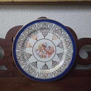 May include: A decorative porcelain plate with a blue rim and gold trim. The plate features a central image of cherubs and ornate gold and black patterns around the edge. The plate is set on a dark wooden surface.