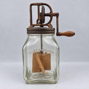 Puede incluir: Una mantequera vintage Dazey-Churn No 40 con un frasco de vidrio cuadrado transparente con letras en relieve, una tapa de metal oxidado marrón oscuro, un mecanismo de engranajes, una manivela de madera y paletas mezcladoras de madera.