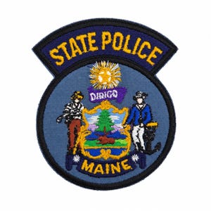 Pode incluir: Um emblema bordado circular com o brasão da Polícia Estadual do Maine. O design inclui um fundo azul, um escudo central com um pinheiro e um alce, duas figuras, um sol radiante e o texto STATE POLICE e MAINE em fio de cor amarela.