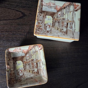 May include: Two square ceramic ashtrays with a detailed street scene. The ashtrays feature a cream-colored base with a yellow border and a painted image of a European town with shops and people.