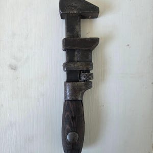 Puede incluir: Una llave inglesa antigua con un mango de madera oscura y una cabeza de metal. La llave está hecha de metal oscuro y envejecido, mostrando signos de desgaste. El mango es de color marrón oscuro.