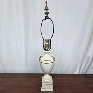 May include: A white ceramic table lamp with gold accents and a black lamp shade holder. The lamp has a decorative urn-shaped base and sits on a square base. The lamp is approximately 24 inches tall.