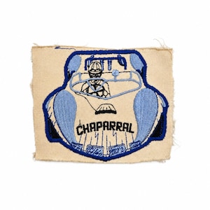 May include: A rectangular embroidered patch on a beige fabric base featuring a blue and black design of a race car with a driver. The car is depicted from a front-facing perspective with the word CHAPARRAL stitched in black thread below the windshield.