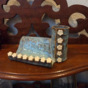 May include: A handcrafted ceramic piece with a blue and white glaze. The piece features a small vase and a sloped tray, both adorned with white flower-shaped accents. It sits on a dark wooden shelf.