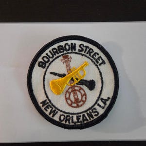May include: Embroidered patch with a white background and black border. The patch features the text "BOURBON STREET NEW ORLEANS LA" in a circular design. A yellow trumpet and a black saxophone are centered above a brown guitar.