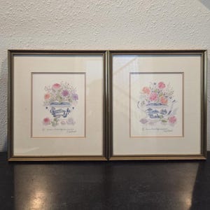 May include: Two framed floral watercolor paintings. Each features a blue and white vase filled with pink, purple, and white flowers. The frames are gold-toned, and the artwork is set against a cream-colored background.