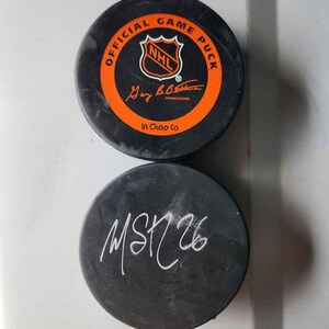May include: Two black hockey pucks. One puck has an orange ring with the text "OFFICIAL GAME PUCK" and the NHL logo. The other puck has the handwritten text "MSK 26" in white.