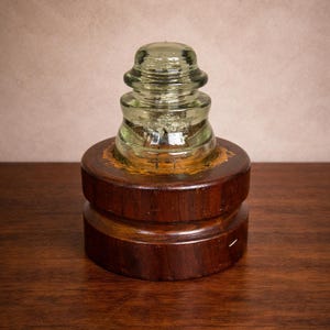 May include: A vintage glass insulator with a layered design, mounted on a dark stained wooden base. The glass has a slight green tint. The wooden base has a two-tiered design. The item is displayed on a dark wood surface.