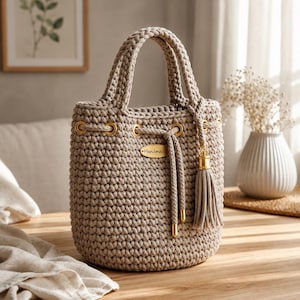 May include: A beige crochet bucket bag featuring a thick braided handle, gold-toned metal grommets, and a drawstring closure with matching cord ends. An oval-shaped wooden tag labeled "Handmade" is attached to the front, along with a tassel.