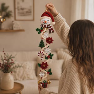 Può includere: Una decorazione natalizia verticale all'uncinetto con un pupazzo di neve in cima che indossa un cappello e una sciarpa rossi. La ghirlanda include elementi lavorati all'uncinetto come stelle di Natale, un pino, bastoncini di zucchero e campanellini.