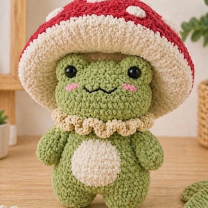 Peut inclure: Une grenouille verte faite au crochet, portant un chapeau champignon rouge et blanc. La grenouille a des yeux noirs, des joues roses et un col couleur crème. Un cercle blanc est sur le ventre de la grenouille. Un jouet en peluche artisanal.