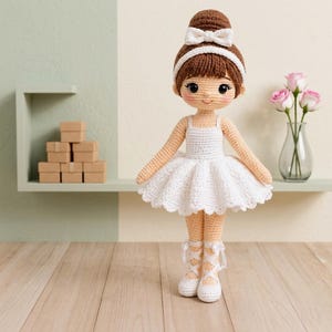 Puede incluir: Muñeca de bailarina de ganchillo hecha a mano, con cabello castaño recogido en un moño y un lazo blanco. La muñeca lleva un tutú blanco, zapatillas de ballet y una diadema. El fondo incluye una estantería con cajas pequeñas y un jarrón con rosas rosas.