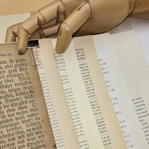 May include: A wooden articulated hand holds several pages of aged paper, secured with a metal binder clip. The pages display text in multiple languages, suggesting a focus on literature or historical documents. The overall aesthetic is vintage.