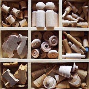 May include: A wooden box with multiple compartments filled with various unfinished wooden toy parts. The parts include spools, peg dolls, wheels, rolling pins, and other shapes. The items are all made of light-colored wood.