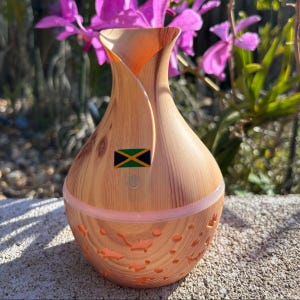 May include: A wood-grain essential oil diffuser with a Jamaican flag emblem. The diffuser has a light pink base with cut-out designs and a power button. The background features purple flowers and sunlight.