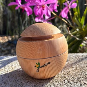 May include: A light brown, wooden essential oil diffuser with a rounded shape and a hinged top. The diffuser has a power button and the word "Jamaica" in green script. The background features purple flowers.