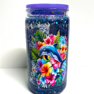 May include: A clear jar with a purple lid filled with blue glitter and a colorful floral and dolphin design. The jar features a vibrant illustration of a dolphin surrounded by tropical flowers in shades of pink, orange, yellow, and blue.