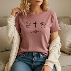May include: A dusky pink t-shirt featuring black graphic icons: a coffee cup, a cross, and a paw print. Beneath these are two hearts joined by a curved line. The shirt is worn with blue jeans and a cream cardigan.