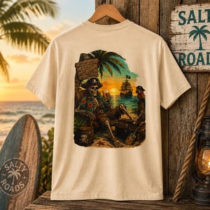 K&ouml;nnte beinhalten: Ein beiges T-Shirt mit einem Grafikdruck eines als Pirat verkleideten Skeletts, das bei Sonnenuntergang in einem Stuhl am Strand sitzt. Das Skelett h&auml;lt ein Glas, im Hintergrund ist ein Piratenschiff und ein Holzschild mit der Aufschrift: Drink like a pirate, live like a legend.