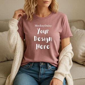 Puede incluir: Camiseta de color rosa polvoriento con el texto "MocksyDaisy Your Design Here" en blanco. La modelo lleva vaqueros azules y una rebeca color crema. La camiseta es de cuello redondo.