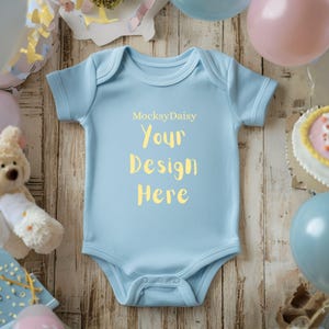Könnte beinhalten: Hellblauer Baby-Body mit gelbem Text "MocksyDaisy Your Design Here". Der Body liegt auf einer Holzoberfläche, umgeben von Luftballons und einem Stofftier.
