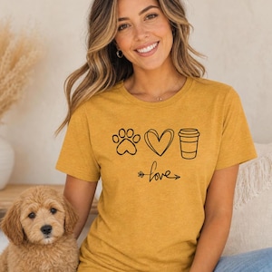 May include: Mustard yellow t-shirt with a black paw print, heart, and coffee cup design. The word "love" is written below the symbols with an arrow. The shirt is made of a soft material.