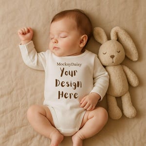Könnte beinhalten: Ein schlafendes Baby in einem cremefarbenen Body mit dem Text "MocksyDaisy Your Design Here". Ein beiges Stoffkaninchen liegt neben dem Baby. Das Baby liegt auf einer beigen Oberfläche.