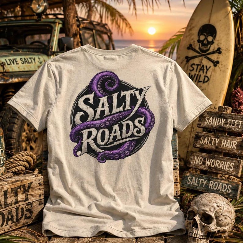 May include: A beige short-sleeve t-shirt featuring a circular graphic on the back. The design includes a purple octopus tentacle wrapped around the text Salty Roads in a stylized font. The shirt is displayed against a beach-themed background.