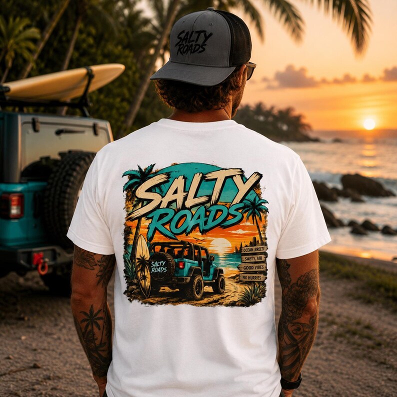 May include: A white t-shirt featuring a graphic on the back of a blue off-road vehicle parked on a beach at sunset with palm trees and a surfboard. The text Salty Roads is printed in large letters above the vehicle, with smaller signs reading Ocean Breezy, Salty Air, Good Vibes, and No Hurries.