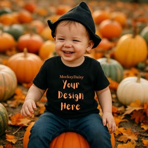 Könnte beinhalten: Ein lächelndes Kind trägt einen schwarzen Hut und ein schwarzes T-Shirt mit dem Text "MocksyDaisy Your Design Here" in Orange. Das Kind sitzt auf einem Kürbis, umgeben von anderen Kürbissen und Herbstlaub.