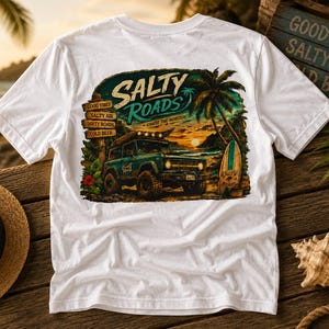 May include: A white t-shirt featuring a graphic print of a teal off-road vehicle on a beach at sunset. The design includes palm trees, a surfboard, and a wooden signpost with the text: SALTY ROADS, CHASE THE HORIZON, GOOD VIBES, SALTY AIR, DIRTY ROADS, COLD BEER.