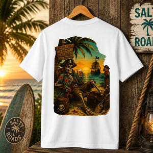 K&ouml;nnte beinhalten: Ein wei&szlig;es T-Shirt mit einem Grafikdruck eines Skeletts mit Piratenhut und Blumenhemd, das bei Sonnenuntergang in einem Stuhl am Strand sitzt. Das Skelett h&auml;lt ein Bierglas, daneben sind ein Schiff, ein Papagei, ein Fass und ein Schild mit der Aufschrift DRINK LIKE A PIRATE LIVE LIKE A LEGEND zu sehen.