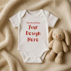 Könnte beinhalten: Ein weißer Baby-Body mit dem Text "MocksyDaisy Your Design Here" in Rot. Ein beiges Stoffkaninchen liegt neben dem Body. Der Body hat kurze Ärmel und Druckknöpfe am unteren Ende.