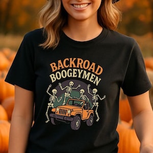 May include: Black t-shirt with the text "BACKROAD BOOGEYMEN" in orange and white. The graphic features skeletons dancing around an orange jeep. The background is a blurred image of pumpkins.