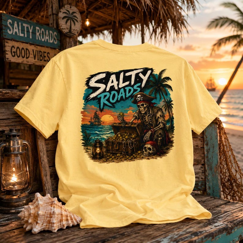 Puede incluir: Una camiseta de color amarillo p&aacute;lido con un estampado gr&aacute;fico de un esqueleto pirata arrodillado junto a un cofre del tesoro en una playa. El dise&ntilde;o incluye palmeras, un barco en el mar, un farol y el texto Salty Roads en una fuente estilizada.