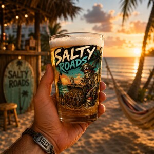 May include: A clear glass filled with a golden beverage, featuring a printed graphic of a pirate skeleton with a treasure chest and the text Salty Roads. The design includes vibrant colors like teal, orange, and yellow, depicting a beach scene.