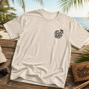Może przedstawiać: Kremowy t-shirt z kr&oacute;tkim rękawem z małą czarną grafiką na lewej piersi z napisem Salty Roads i ikoną palmy. To samo logo znajduje się na pasującej czapce z daszkiem oraz na prostokątnej drewnianej tabliczce obok koszulki.